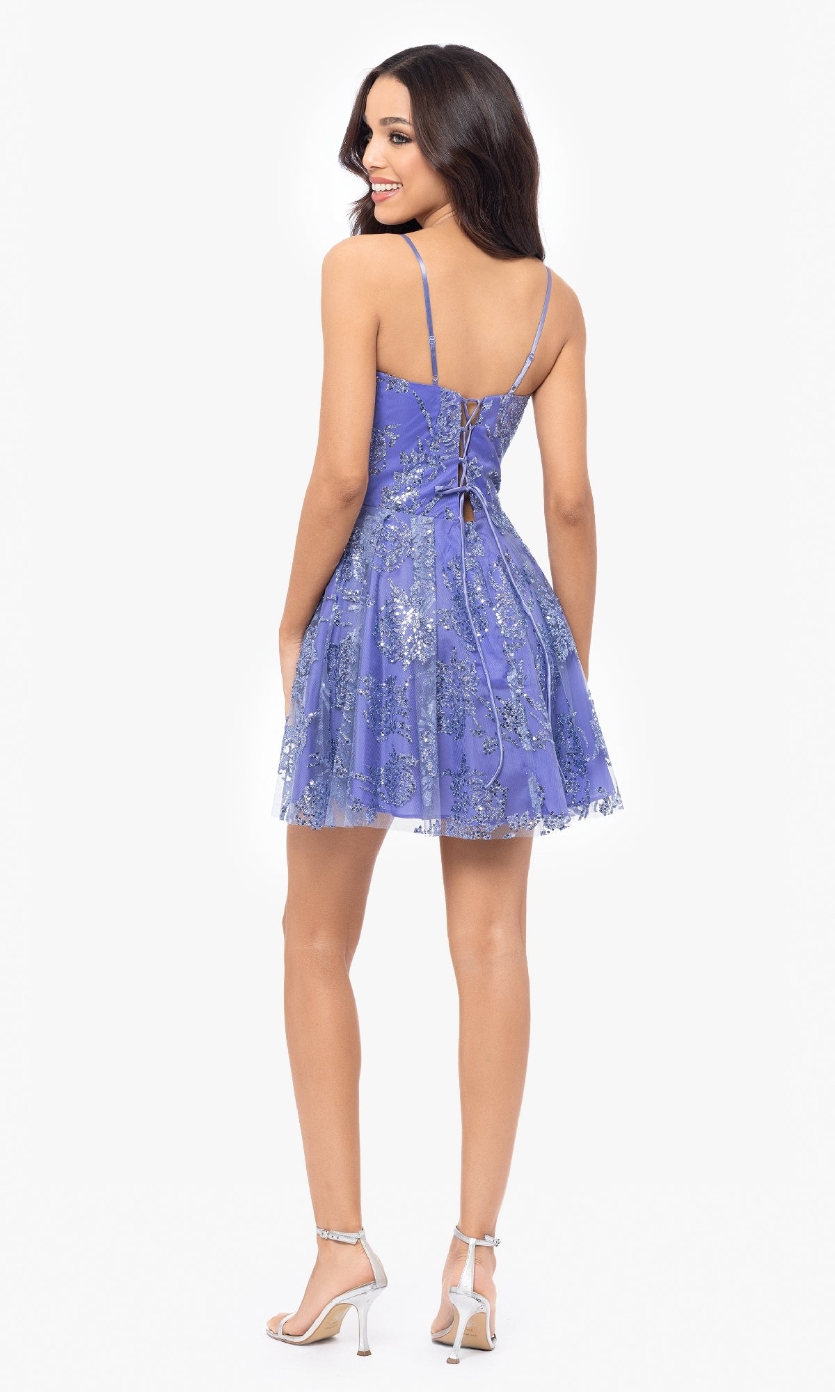 A woman in the Short Periwinkle Hoco Dress: Blondie Nites 5017BN, featuring silver floral patterns and a lace-up back, stands smiling and facing away from the camera.